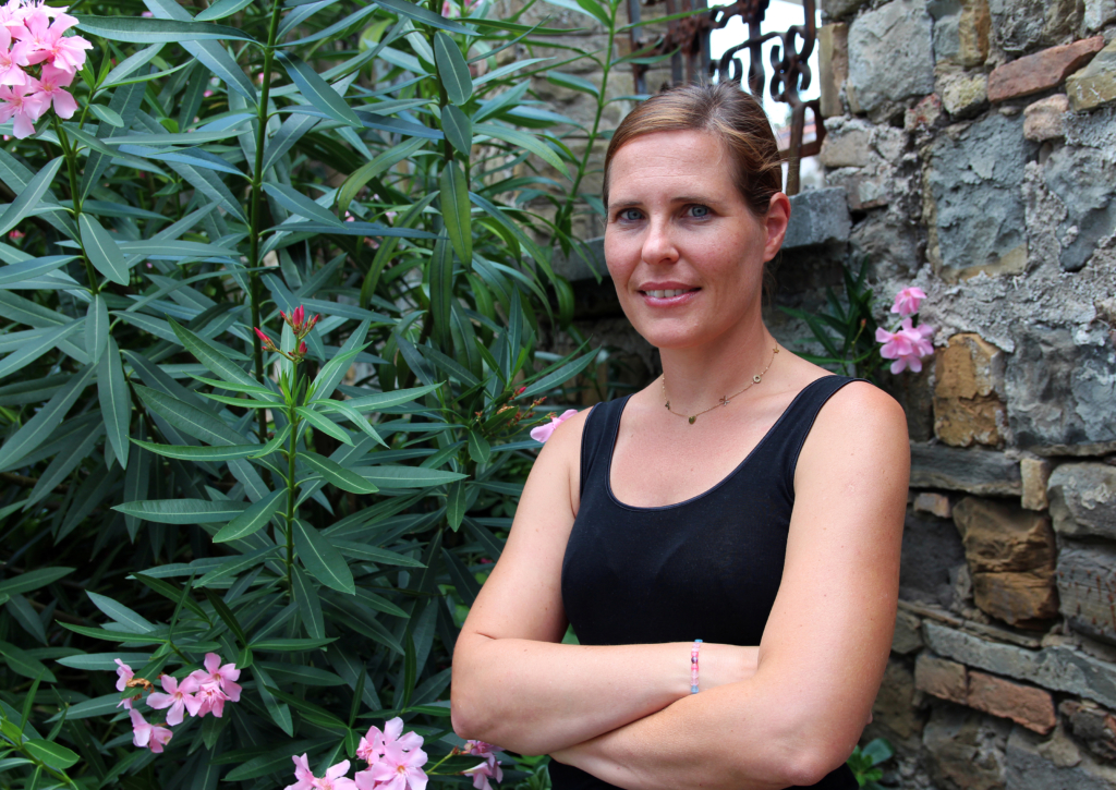 Portrait photo of a women with her arms folded wearing a black vest standing outside infront of a large green and pink plant