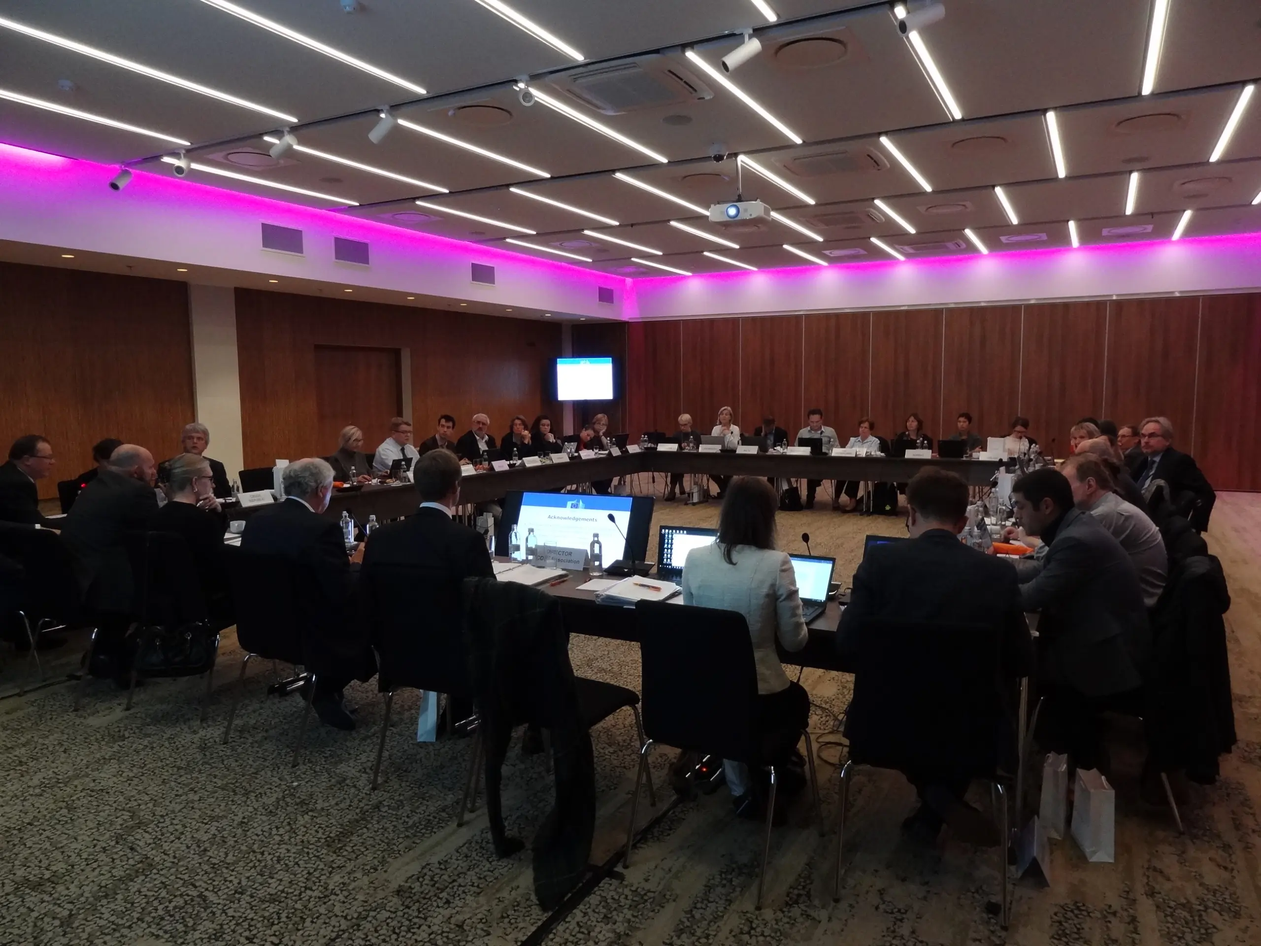 Photo of a business meeting with around 50 participants sitting aroung a large square table in discussion with a large screen in the far left corner