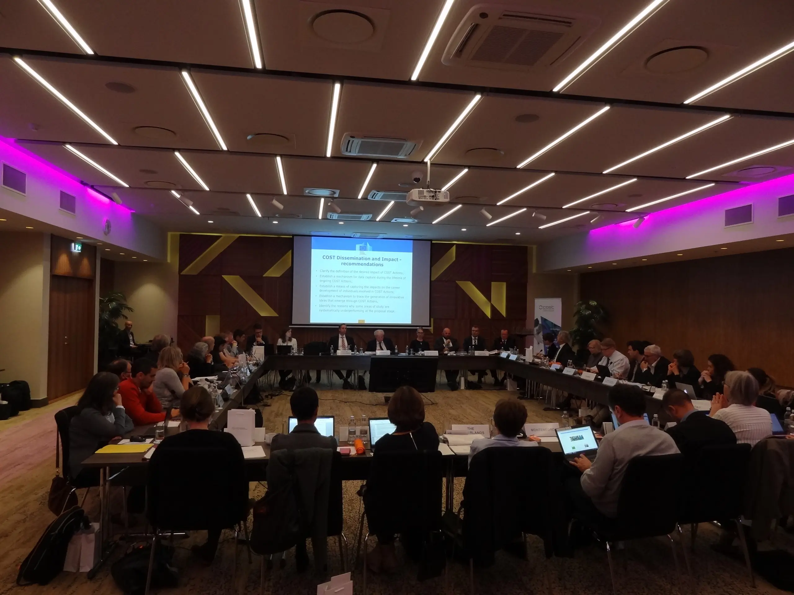 Photo of a business meeting with around 50 participants sitting aroung a large square table in discussion with a large projection screen on the back wall of the room