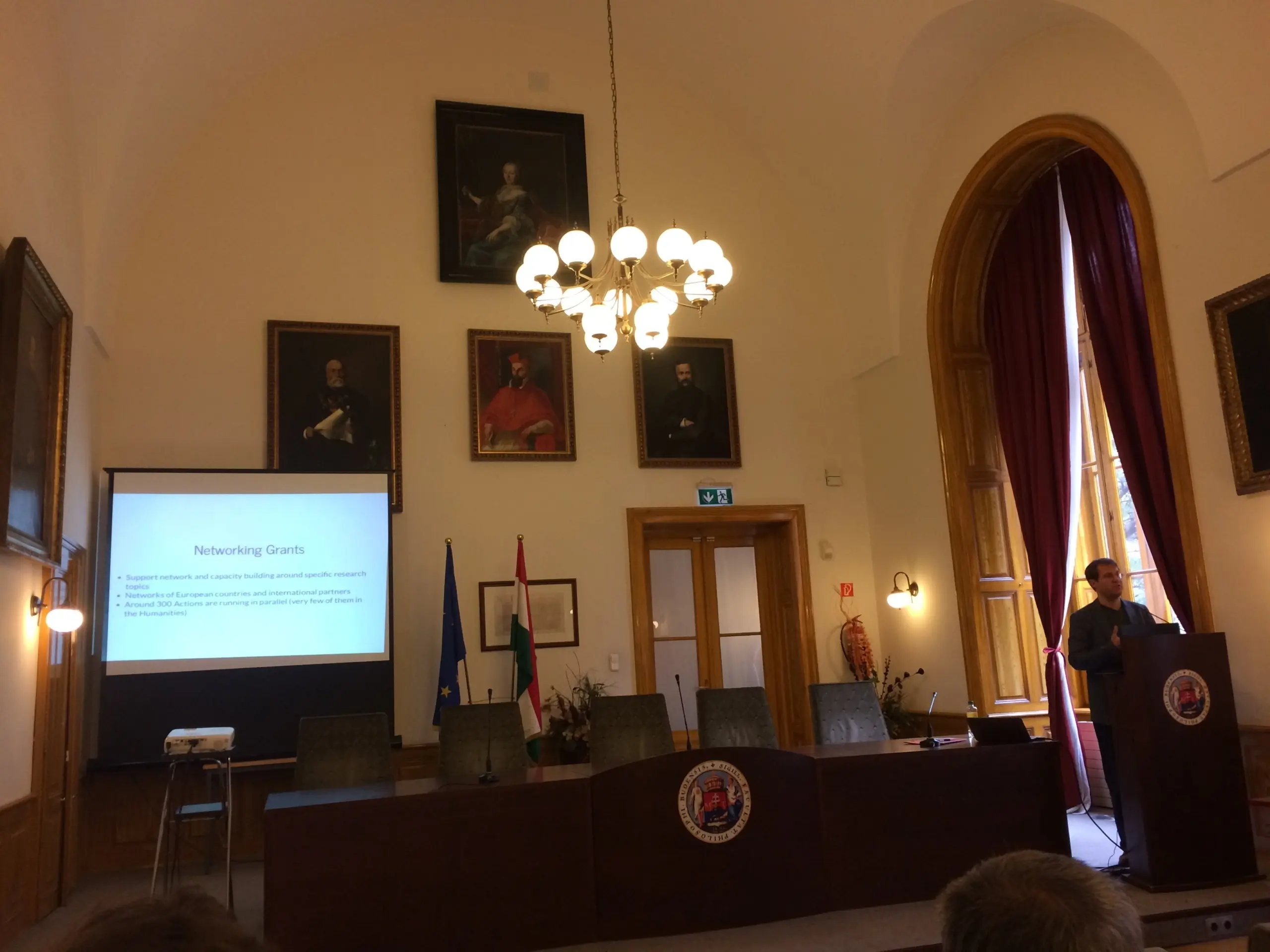 Photo of a grand room with very high ceilings and paintings on the wall with a man standing at a lectern giving a presentation