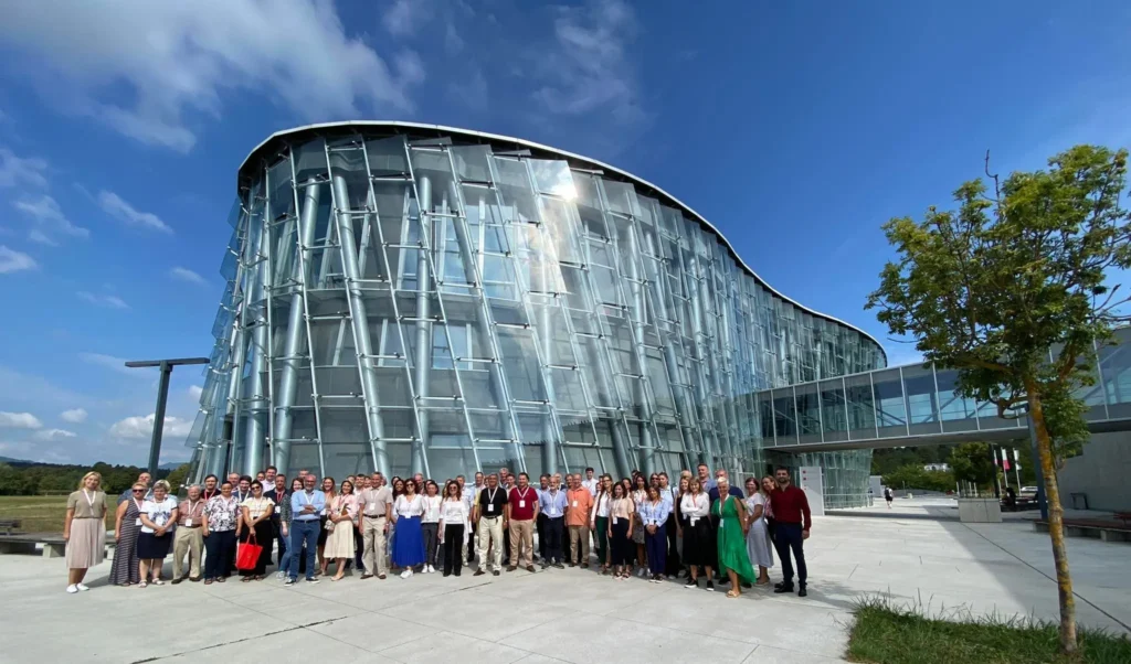 Group of 100+ conference attendees standing outside a modern glass fronted building posing for a group photo on a sunny summer day