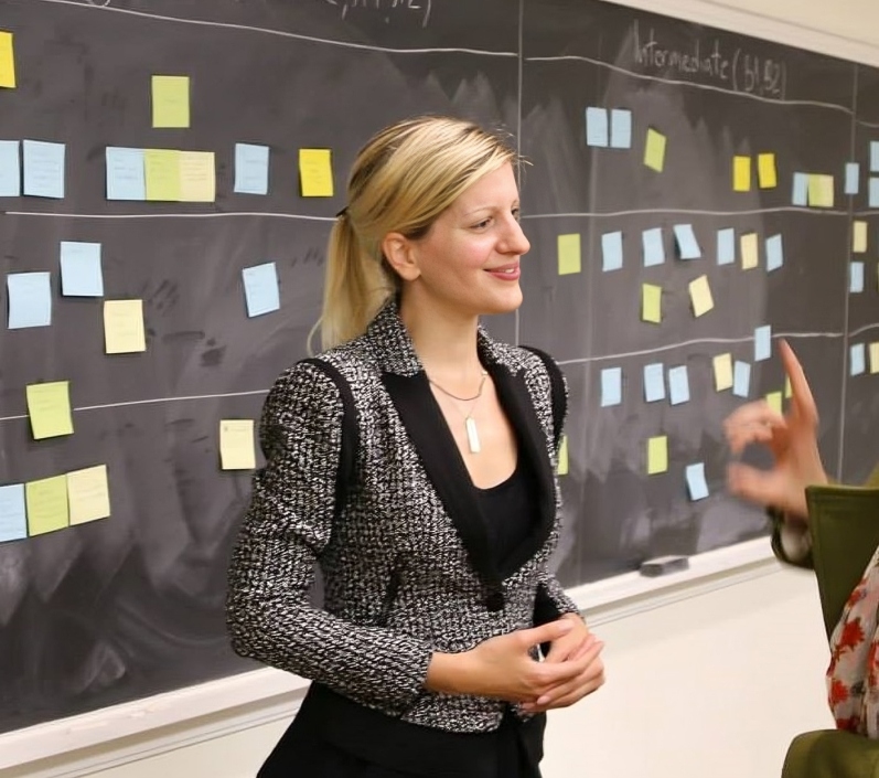 Photo of a young blonde woman standing in front of a black bord.
