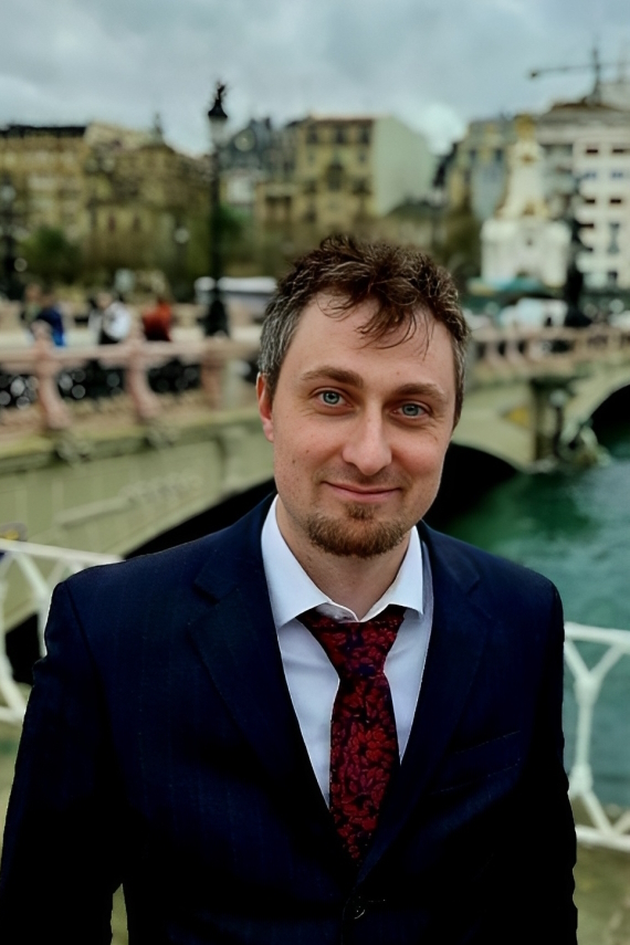 Photo of a smiling young man dressed in a dark suit and a tie, standing on a bridge.