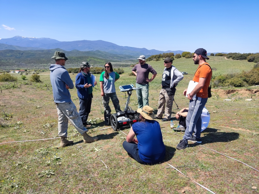 A group of people are positioned in a circle in a field, with some equipment on the ground.