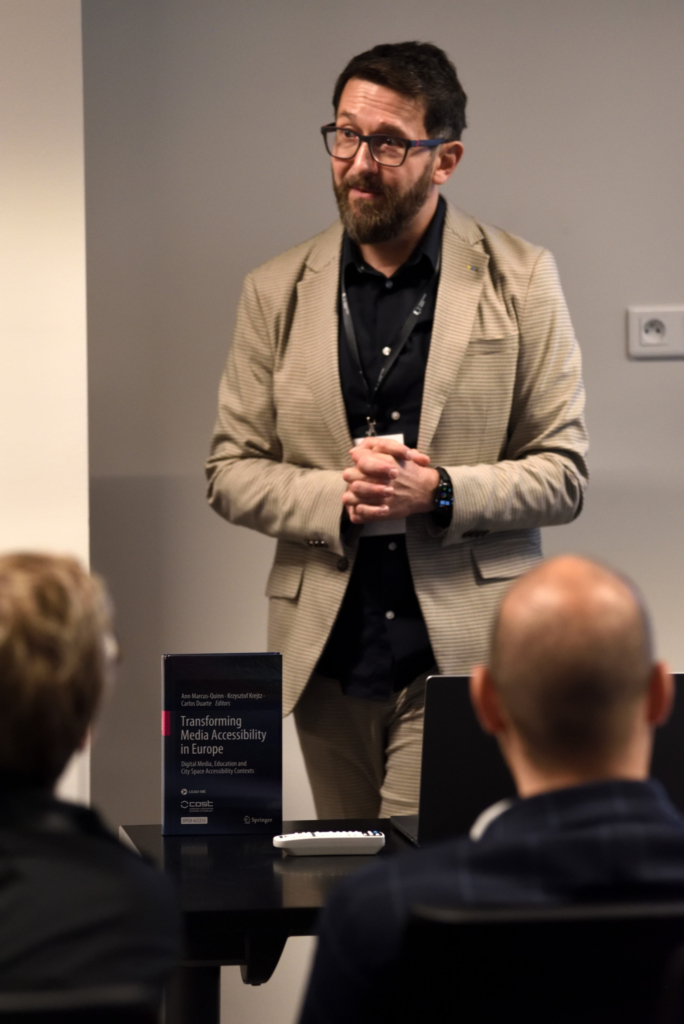 Middle aged man, Krzysztof Krejtz, with beard in a plaid suit wearing black glasses looks pensive during a presentation