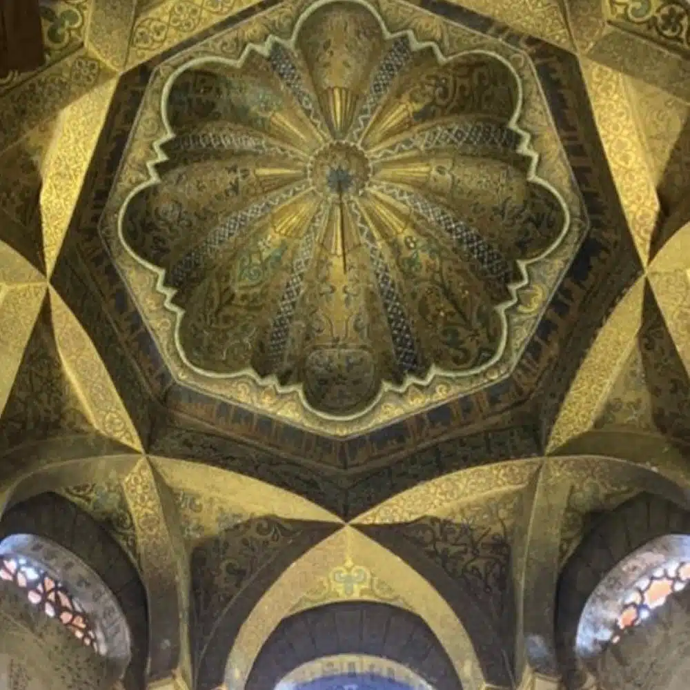This image shows an ornate ceiling inside a mosque, designed in a flower-like pattern with rich gold and green tones. The dome is shaped like a blossoming flower with twelve rounded petals, each decorated with intricate floral and geometric patterns. The surrounding arches and walls are also covered in detailed designs, creating a feeling of elegance and sacred beauty.
