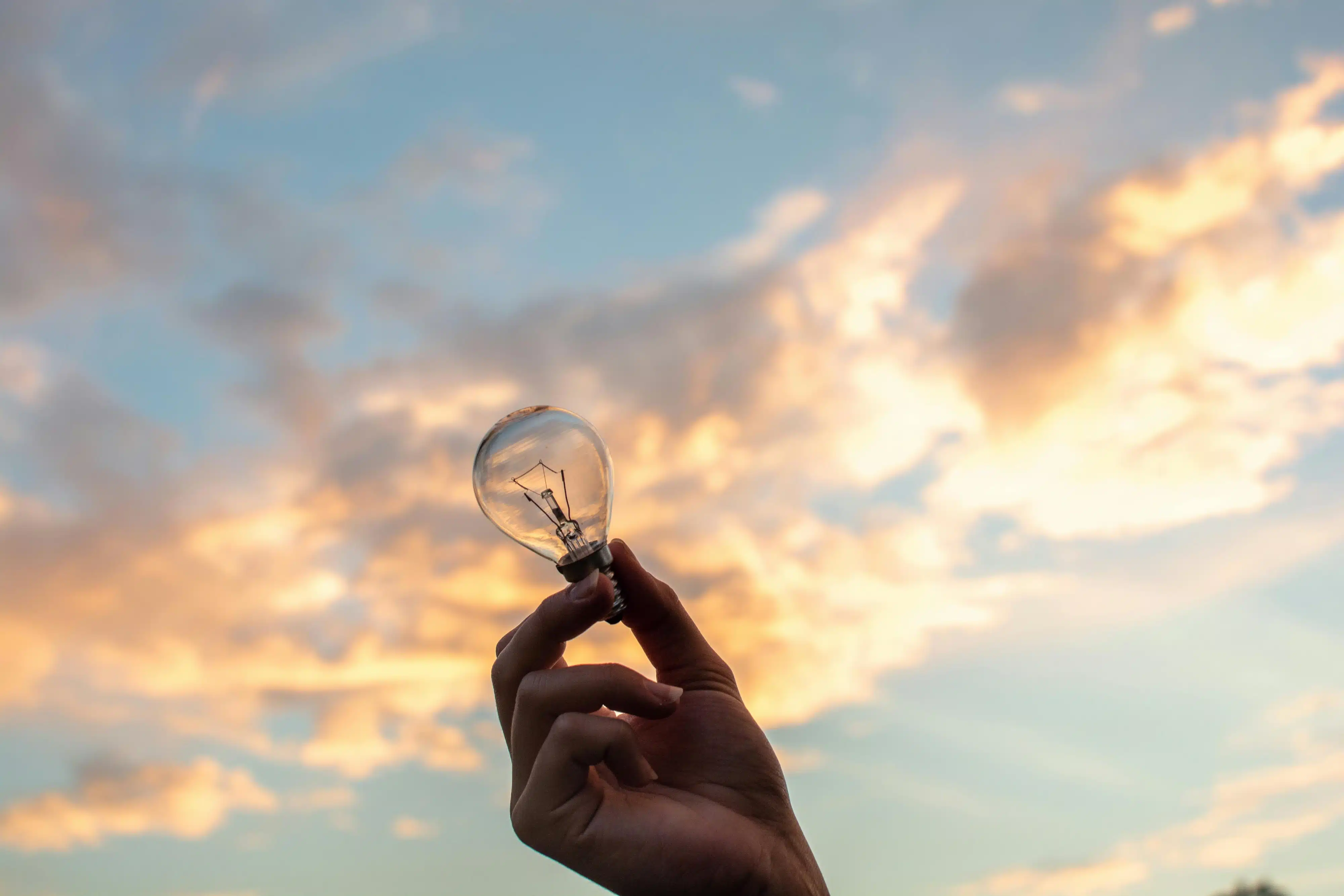 A hand holding a lightbulb up to a cloudy blue sky